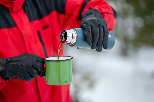 Close-up of hiker pouring hot drink from thermos into travel mug in the snowy forest