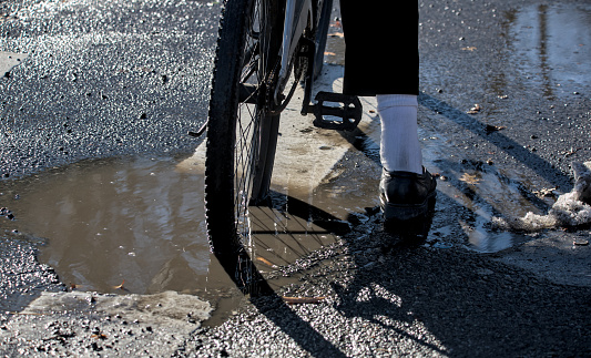 bicycle rear wheel in deep pothole filled with dirty water during winter (road construction, bike stuck inside hole) street streets crosswalk (shoe sock pants) detail close up bad conditions