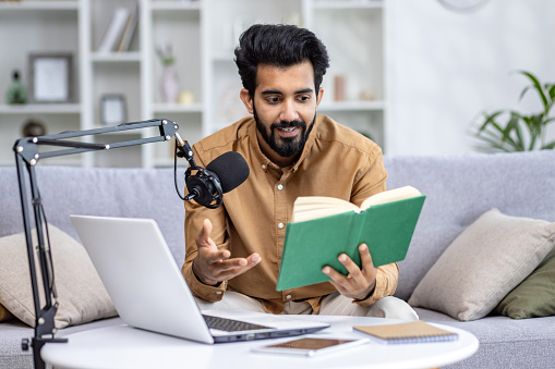 Indian smiling young man sitting on sofa at home in front of laptop and desk with microphone, reading book, online lecture, recording webinar.
