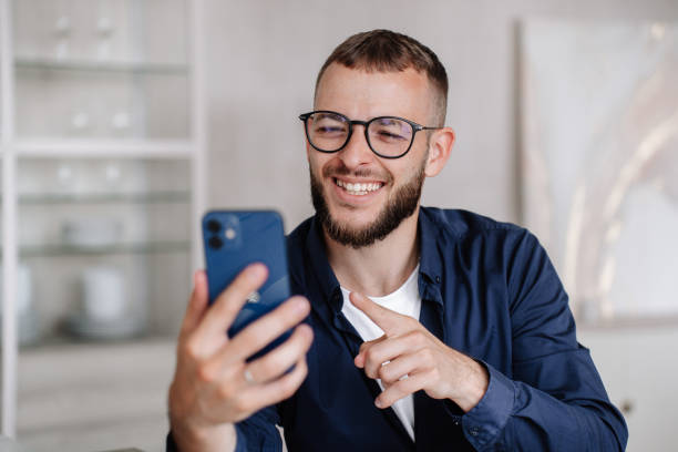 istock Handsome caucasian beardy young man in glasses using phone sits at desk makes video call toothy smiles points at screen reacts on joke. Successful Italian architect discussing with wife via internet. 2033142717