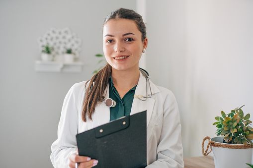 Photo of a confident female doctor holding a clipboard in her home. A plant in the background.