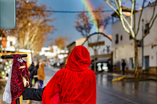 Castilla la Mancha, Spain. Rear view of a woman in a red velvet robe and hood, on a rain-soaked village street, looking at the rainbow on a rainy afternoon.