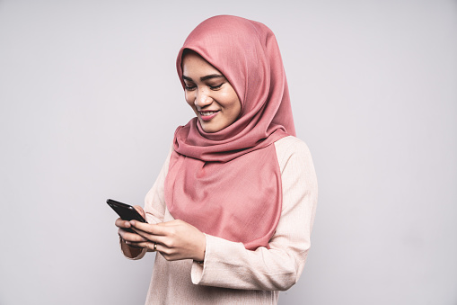 Happy smiling Malaysian Woman standing in Studio, looking down to her Smart Phone with a happy natural smile, wearing a fashionable modern pink-colored Hijab - Headscarf. Using her Smart Phone - Mobile Apps Concept, White Background Portrait Studio Shot. Kuala Lumpur, Malaysia.