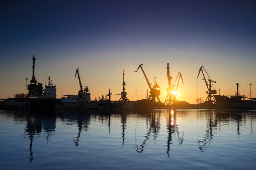 Industrial loads freight at dock. Sunset silhouettes cargo ships, cranes at sea port. Trade, transport infrastructure. Maritime logistics, global shipping. Clear sky reflects calm water.