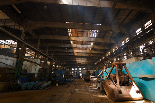 Derelict factory interior, urban exploration, decaying infrastructure, sunlight beams. Abandoned industrial warehouse with rusty machinery, old equipment scattered, sunlight piercing through roof.