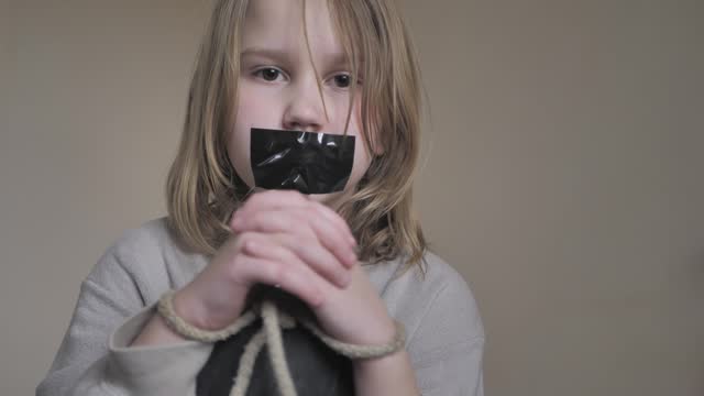 Portrait of a teenage girl with duct tape glued to her mouth and her hands tied with a rope, sadly looking at the camera. Child protection concept he domestic violence. Captive child