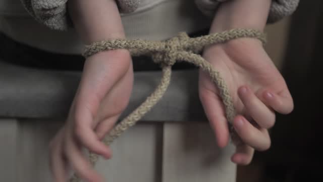 Close-up of a child's small hands tied behind his back with a rope. The child is trying to free his hands from the shackles of the rope. family violence against children. Child in captivity, hostage