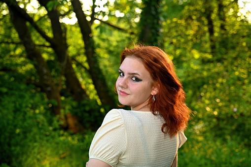 Portrait of a young pretty red-haired girl,  with a light blouse, in green nature, looking at the camera with a smile