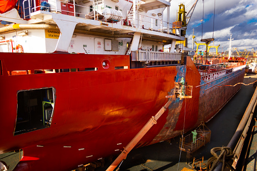 Workers paint, repair vessel hull. Ship undergoing maintenance in floating dock. Industrial, commercial shipyard activity during daytime. Maritime, nautical, shipbuilding business backdrop.