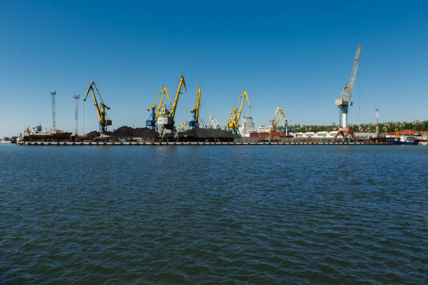 Unused loading equipment stands against clear sky, hinting at past maritime commerce vitality, awaiting repurpose or dismantle. Derelict port cranes line harbor, symbolizing industrial decline.