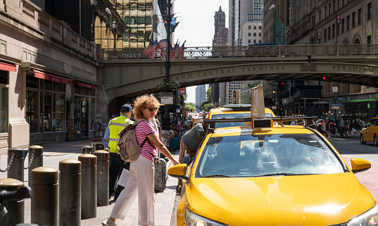 Woman hailing a yellow taxi on a busy street by Grand Central Terminal with American flags in the background