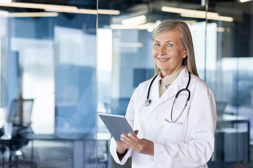 Portrait of a smiling senior female doctor standing in a clinic in a white coat with a stethoscope, holding a tablet and looking at the camera.