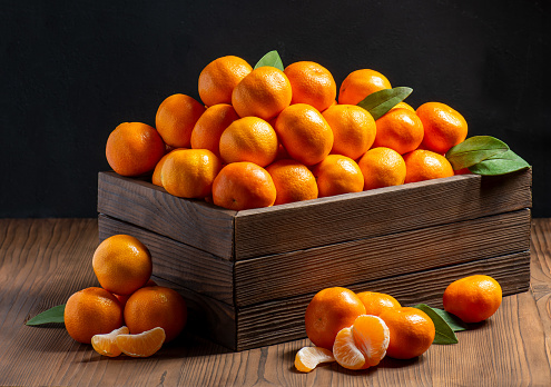 A group of orange ripe tangerines with peeled segments and green leaves lie in a wooden box on a dark table
