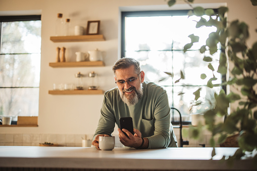 Mature man standing alone in kitchen, drinking coffee and using smart phone.