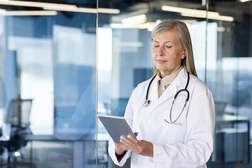 Serious senior gray-haired female doctor standing in clinic and using tablet. Close-up photo.