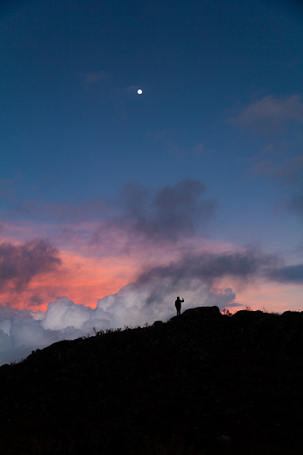 silhouette of a person taking a photograph or selfie in mountain ridge at dawn or dusk with full moon