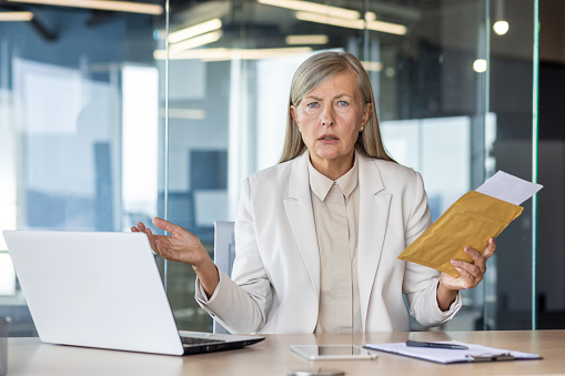 Portrait of an upset and disappointed senior business woman working in the office, sitting at the table, holding a letter in her hands and spreading her hands.