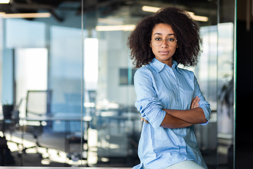 Portrait of a confident African-American young business woman sitting in the office at the desk, hands crossed and looking seriously at the camera.