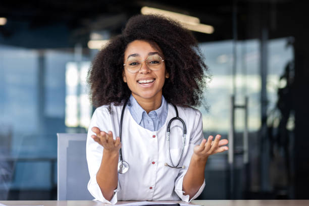 istock Successful female surgeon in professional uniform raising hands while sitting by desk with toothy smile. Positive black woman ready announcing good news and positive prognosis of treatment results 2033739630
