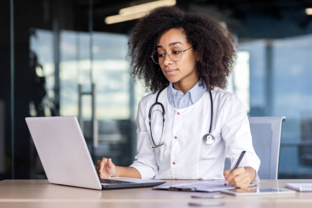 istock Concentrated young African American female doctor, student practitioner studying and working online on laptop, sitting at desk in office and taking notes 2033755082