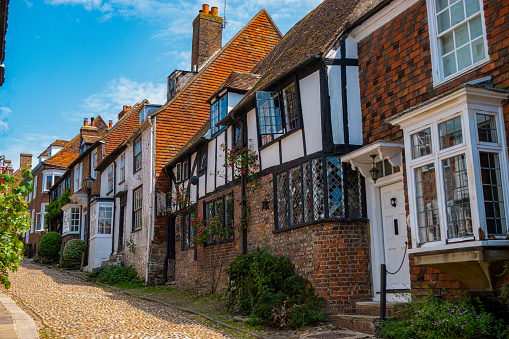 Delightful cobbled street with old timbered and brick houses lined with flowers. Exploring through medieval town centre of Rye. Picturesque fairytale streets in historic cities across southern England