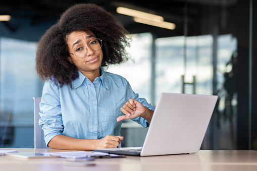 Upset black woman in blue shirt and eyewear making dislike gesture while working by desktop in office. Displeased female manager feeling bored during busy day and dreaming about changing position.
