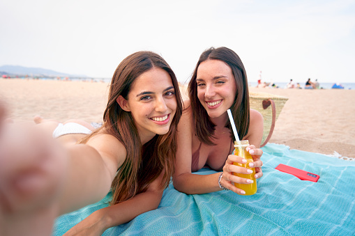 Selfie portrait of two caucasian women laying down on the beach. These girl friends smile together and look pretty and happy. They are young attractive and cheerful.