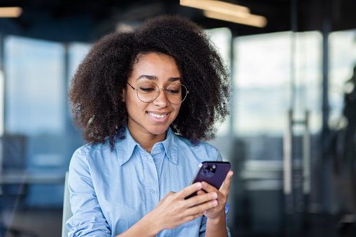Close-up photo of a young smilling African-American woman in an office using a mobile phone.