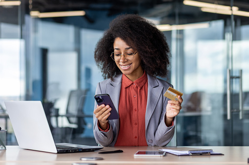 Satisfied african american female keeping debit card and cellular phone in hands by desk with computer and notebook. Pleased lady entering bank data in application for completing cashless payment.