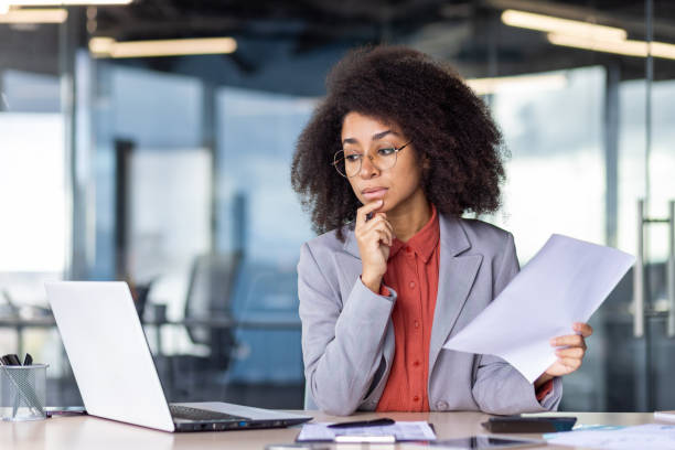 istock Focused project manager with curly hair holding document and looking with hesitation at computer screen. Suspicious woman checking accuracy of printed report and comparing with electronic version 2033857035