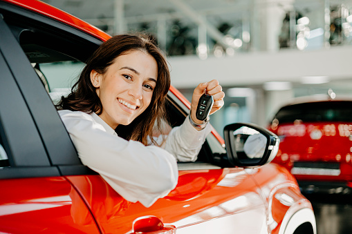 Positioned in her new car with the keys ready, a woman is poised to chart her own course, symbolizing a readiness to face new challenges and embrace the journeys ahead