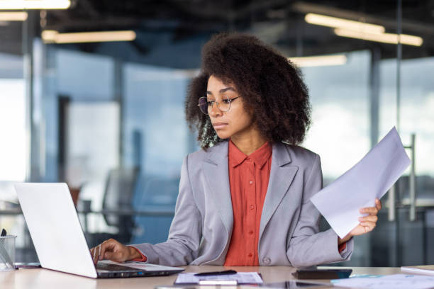 istock Calm diverse lady in formal clothes typing on portable computer keyboard with one hand while keeping sheet of paper. Serious woman making contract for client and printing it in personal workplace 2033882635