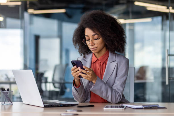 donna d'affari focalizzata con i capelli ricci che utilizza lo smartphone in un moderno ambiente d'ufficio - multi tasking efficiency financial advisor business foto e immagini stock