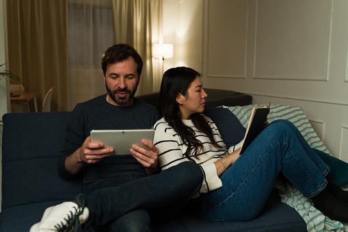 Relaxed married couple using a tablet and reading a book together during their leisure time while relaxing on the couch