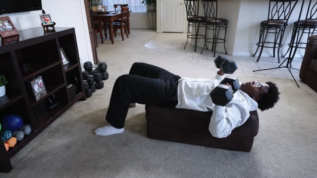 Young African American Man in His Twenties Bench Pressing Dumbbells at Home Using an Ottoman as a Bench to Exercise