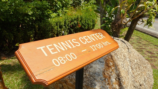 Tennis center information sign displayed on wooden board with operation hours in lush green outdoor setting. Directional signage and sports facility accessibility.