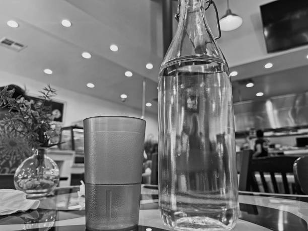 A glass less than half full with water.   A tall glass bottle of water on the table    A small vase with flowers on the left.   Black and white photograph.