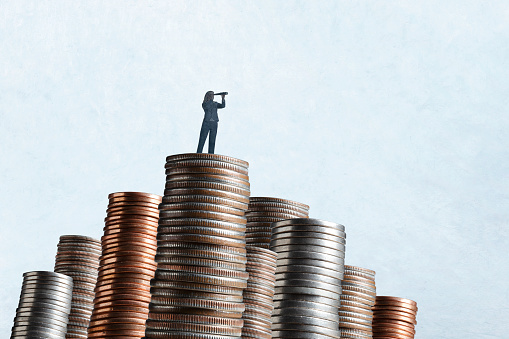 A woman standing on top of a tall stack of coins looks through a spyglass.