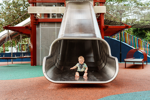 A cute nine month old Eurasian baby boy sits at the bottom of a large metal slide while playing on a playground at a public park in Singapore.