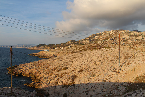 Wooden electric pylon and electric wires in the Calanques of Marseille, Les Goudes, France. Limestone rocks. Beautiful panorama with a cloudy evening sky. Mediterranean sea.