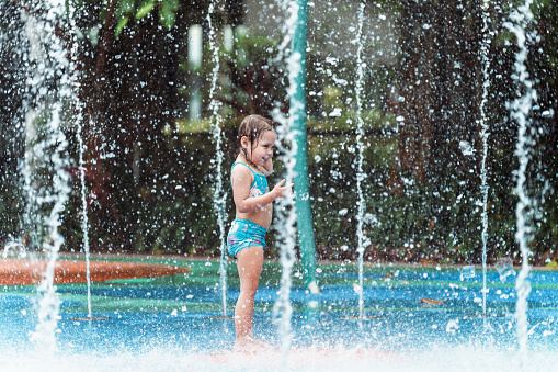 An adorable three year old girl of multiracial descent wearing a swimsuit stands in a fountain of water, playing at a waterpark during a family vacation in Singapore.