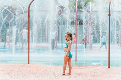 A young girl of multiracial descent looks behind her and smiles while walking through a water fountain and spending the day at a park during a vacation in Singapore.