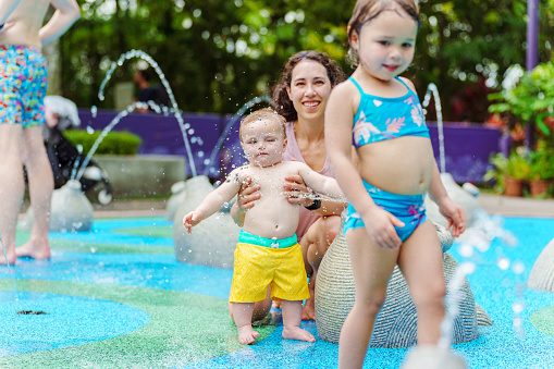 A smiling Eurasian mother sits in a waterpark splash pad, playing with her toddler son and daughter while on a. family vacation in Singapore.