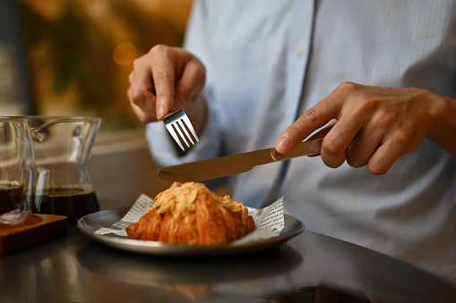 Cropped shot view of man cutting a croissant with a fork and knife, enjoy eating a croissant for breakfast.
