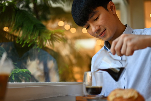 Cropped image of Asian male pouring freshly brewed coffee from pitcher to glass in the cafe.