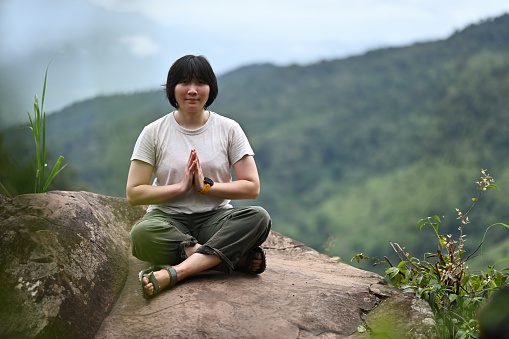 Asian female climber meditating on rocks and natural mountain landscape in the background..