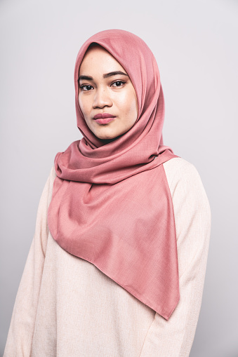 Malaysian Young Woman standing in Studio, looking straight and serious towards the camera, wearing a fashionable modern pinkish colored Hijab - Headscarf. White Background Portrait Studio Shot. Kuala Lumpur, Malaysia.