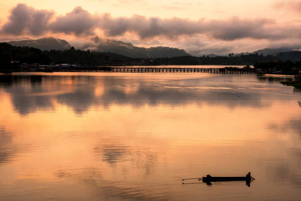 Silhouette fisherman fish on boat by wood Mon bridge with light reflection at dawn on Songkalia river, Sangkhlaburi, Kanchanaburi, Thailand. Famous travel destination landmark or holiday maker in Siam
