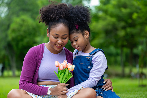 African mother is holding bouquet of tulip flowers and the daughter hugging each other while happily enjoying picnic in the public park during summer for family love and care in mother's day