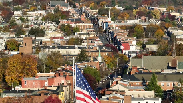 American flag waving in American city during autumn. Aerial rising shot with beautiful urban cityscape background.
